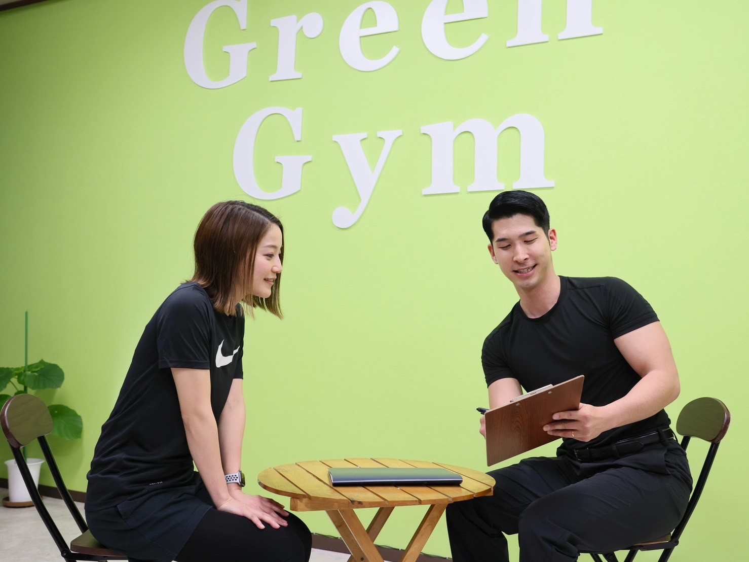 GreenGymのロゴの壁の前で、トレーナーが女性のお客様にカウンセリングをしている様子