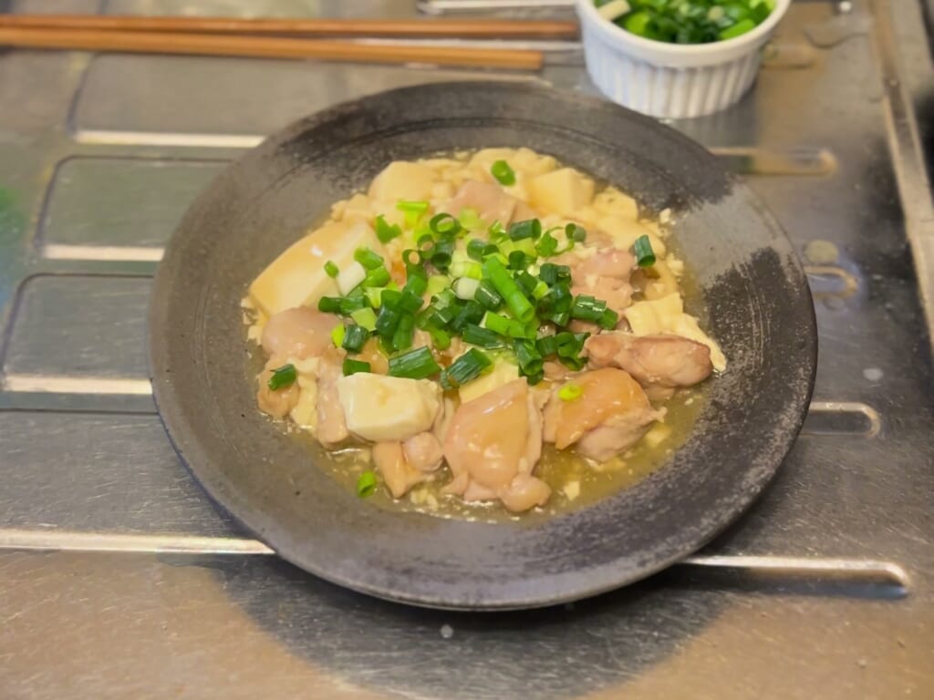 黒いお皿に盛った鶏もも肉と豆腐の和風あんかけ。刻みねぎをのせ、奥に刻みねぎの小鉢